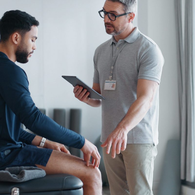 Physiotherapy, tablet and knee pain with people in clinic for rehabilitation, healthcare or consulting. Support, medical and chiropractor with man and athlete for healing, advice or physical therapy.